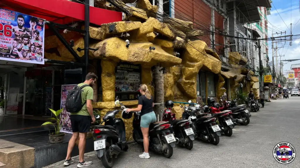 Tourists standing by parked scooters outside a Phuket Muay Thai stadium with yellow stone decor