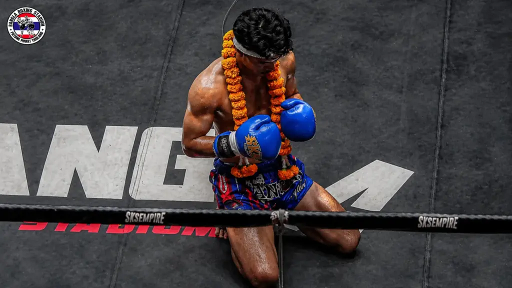 Muay Thai fighter kneeling in prayer with blue gloves and traditional floral garland at a stadium.