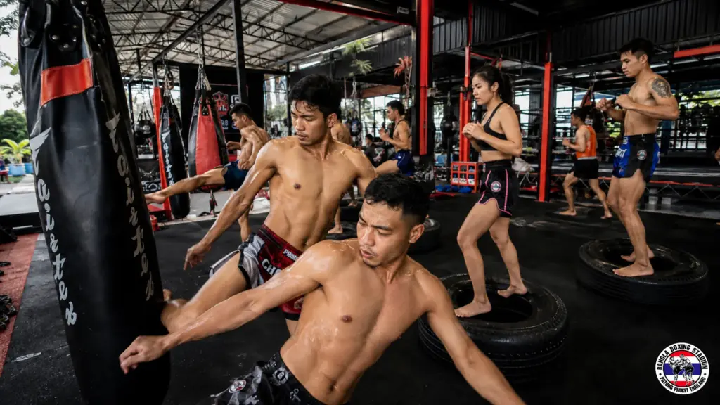 Professional Muay Thai fighters training with heavy bags and tires at a gym in Thailand.