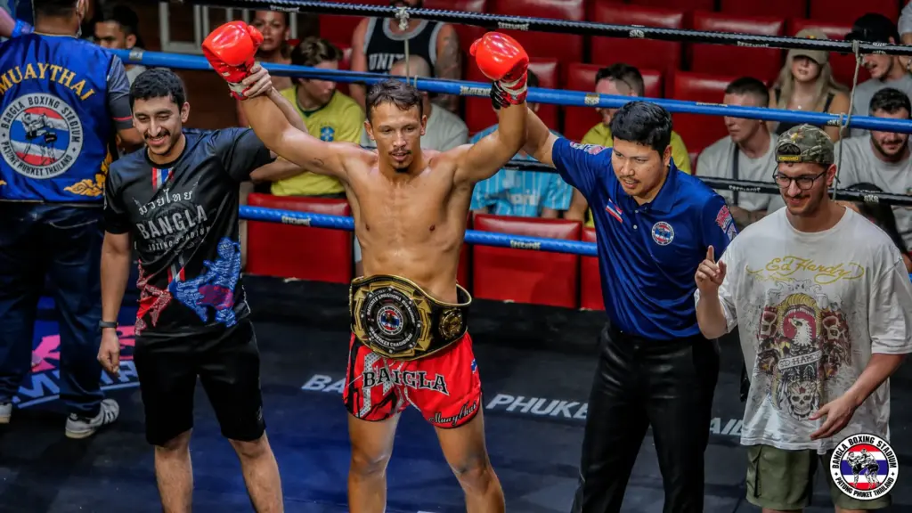 Muay Thai live winner celebrating with championship belt and red gloves at the stadium