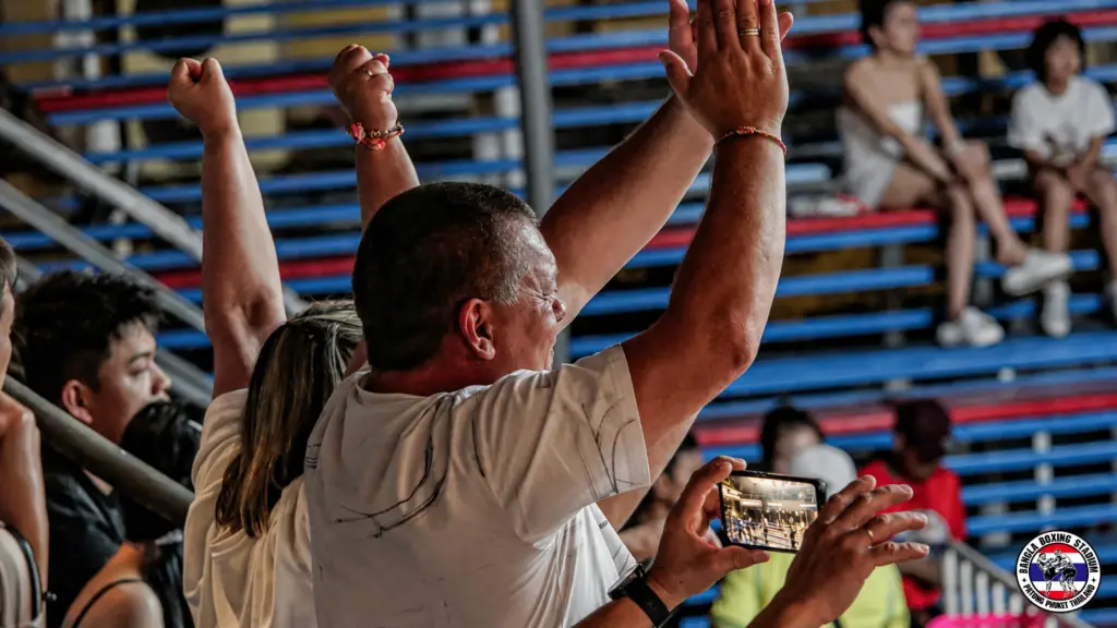 Enthusiastic fans cheering and filming muay thai live matches from the stadium bleachers.