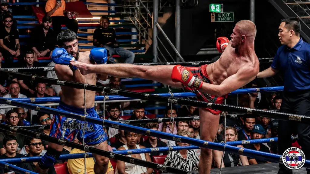 Muay Thai fighter in red shorts delivers a high kick to an opponent in a live boxing ring match.