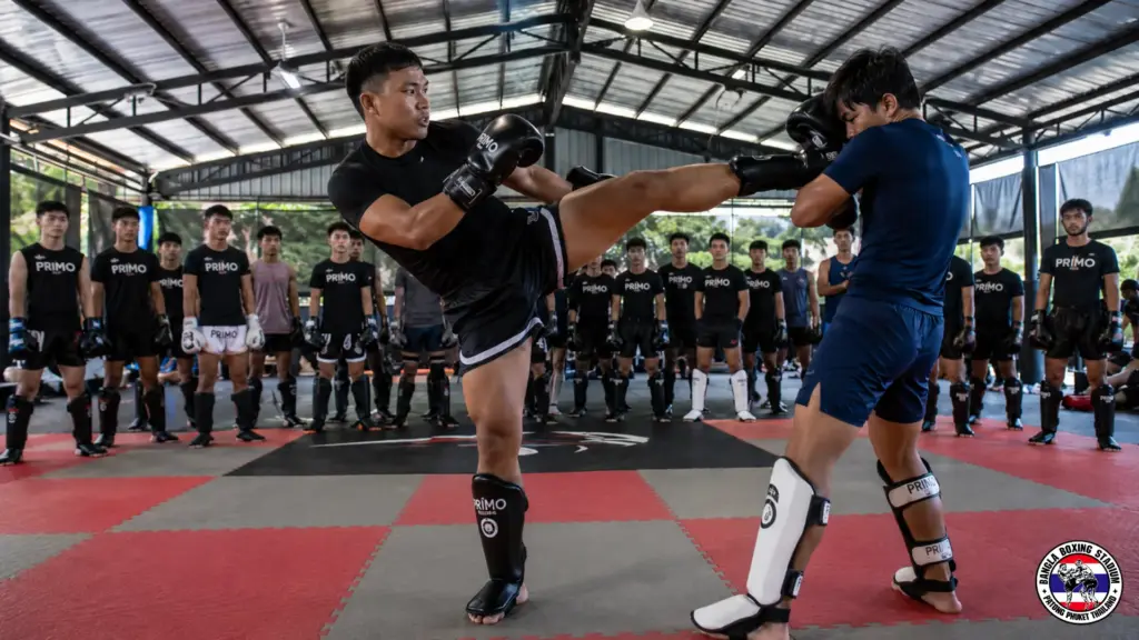 Muay Thai athletes practicing high kicks during training holidays in Thailand gym setting.